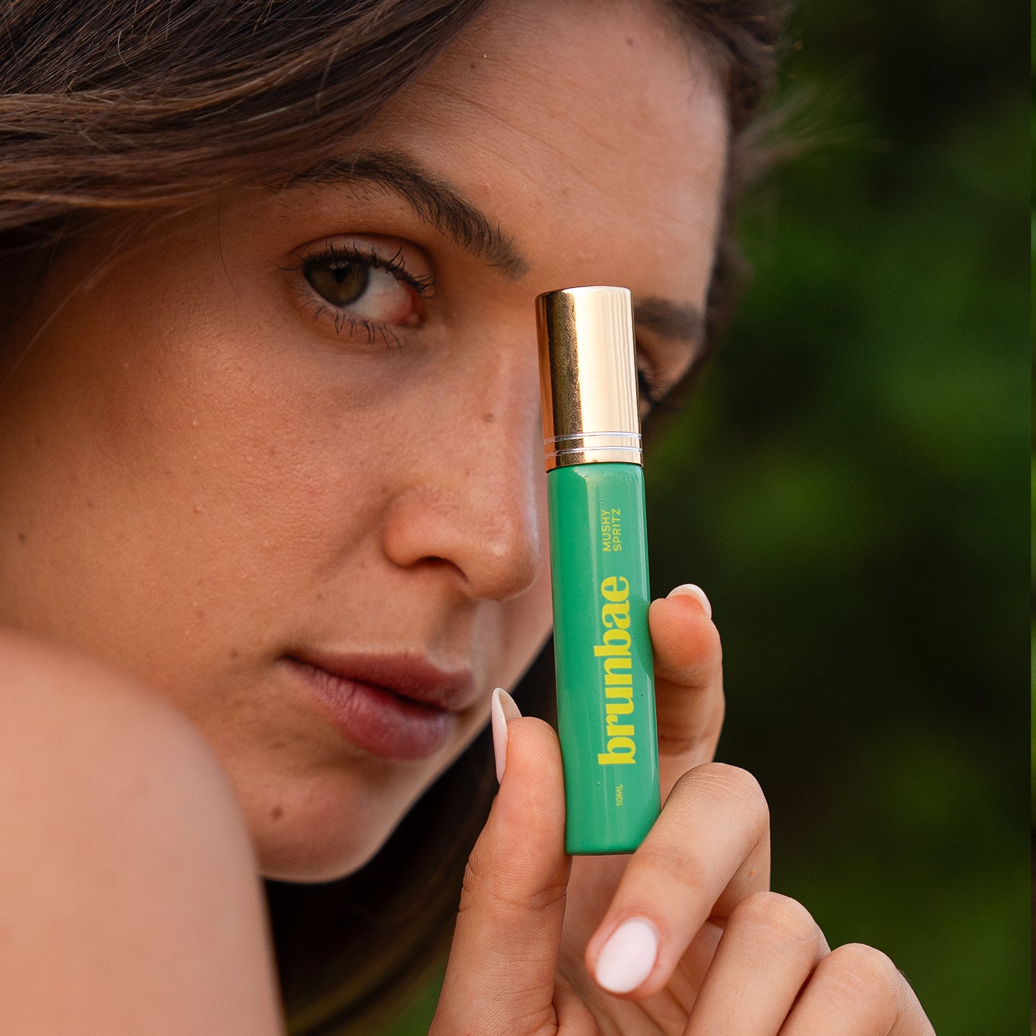 Woman holding a green and gold Brunbae Mushy Spritz (10ml) against her face with a blurred green background
